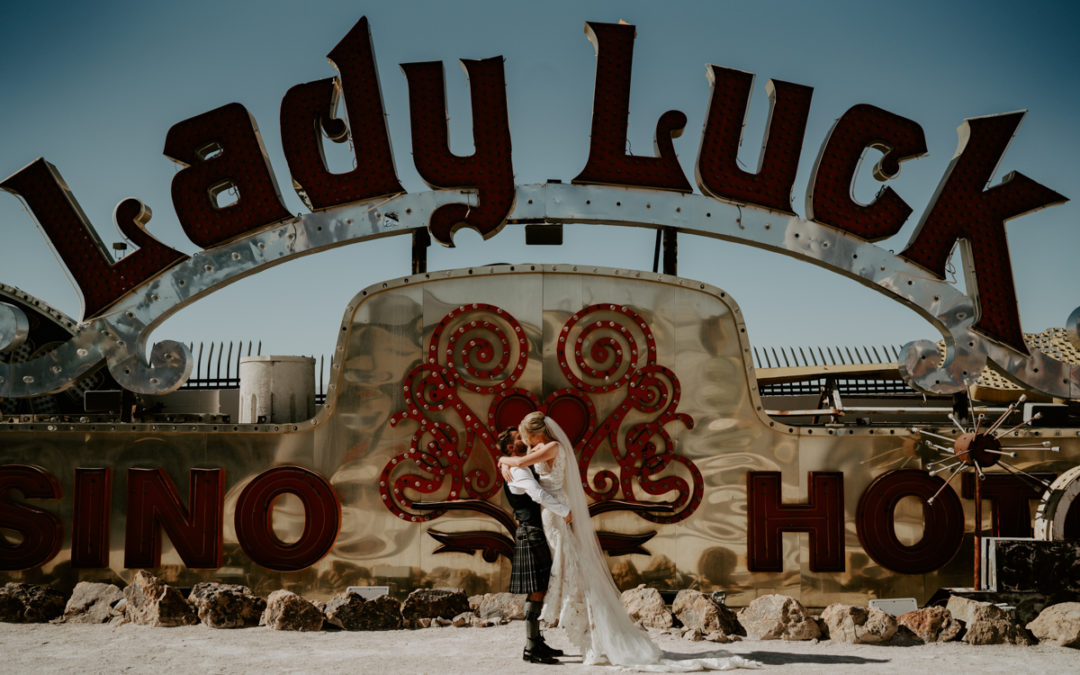 A bride and groom embrace in the desert in front of a vintage vegas neon sign reading lady luck.