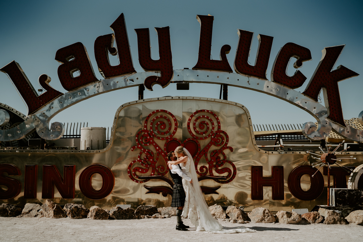 A bride and groom embrace in the desert in front of a vintage vegas neon sign reading lady luck.