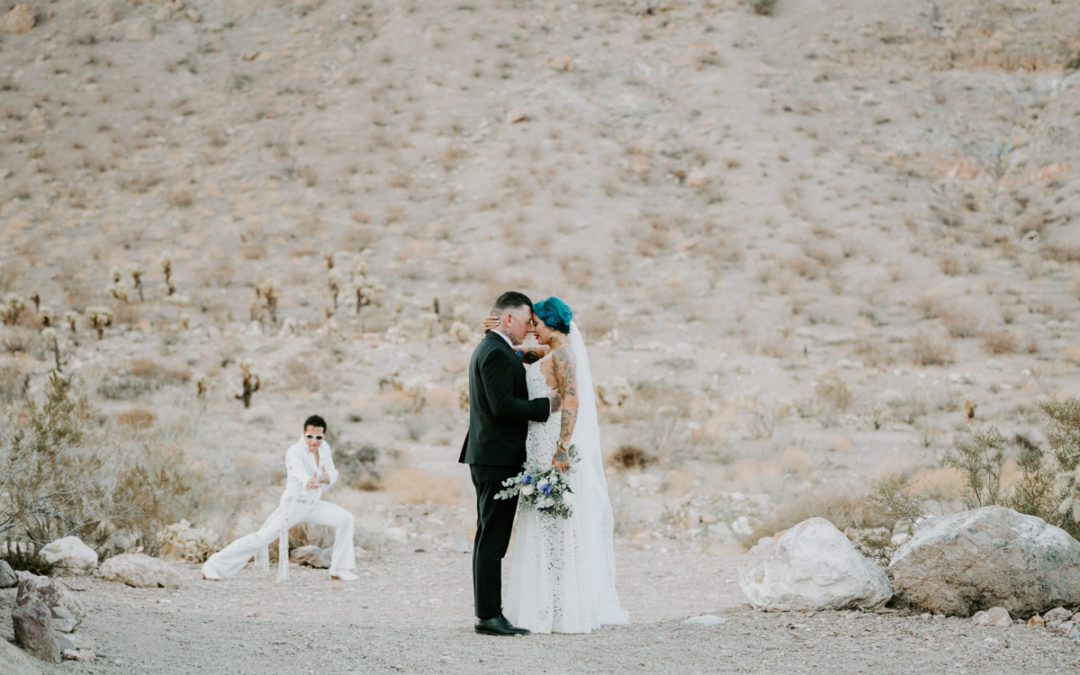A groom and bride press their foreheads together and embrace in the desert with an Elvis impersonator posing in the background.