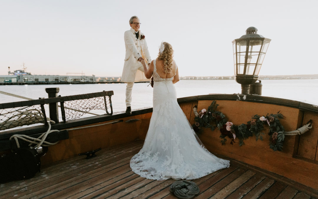 Groom wearing 18th century style white suit stands on the side of a boat and holds his bride's hand and smiles down on her. Her back is to the camera and she's wearing a long white dress.