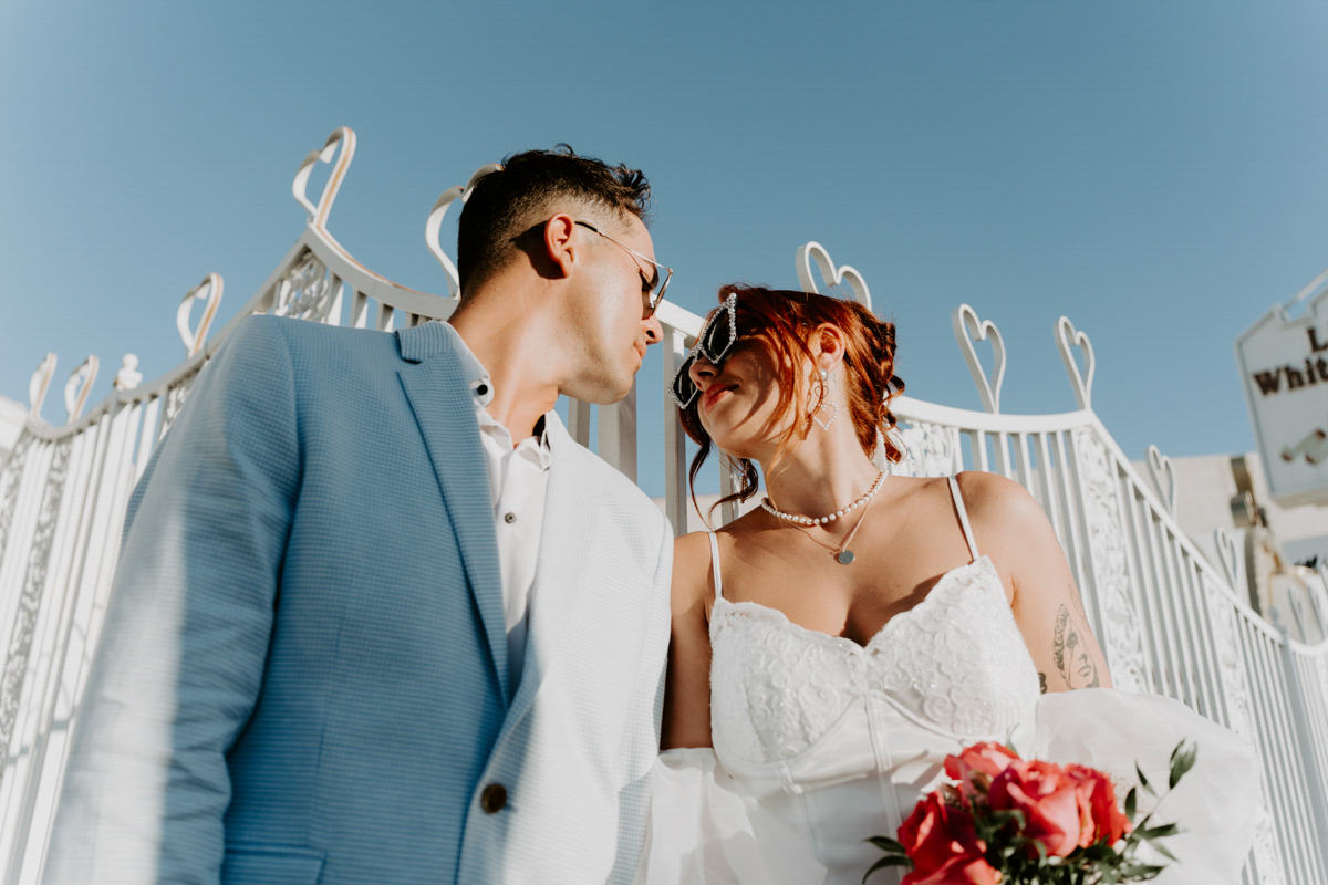 Groom and bride looking at each other, both wearing sunglasses. The photo is taken from below with blue sky in the background.