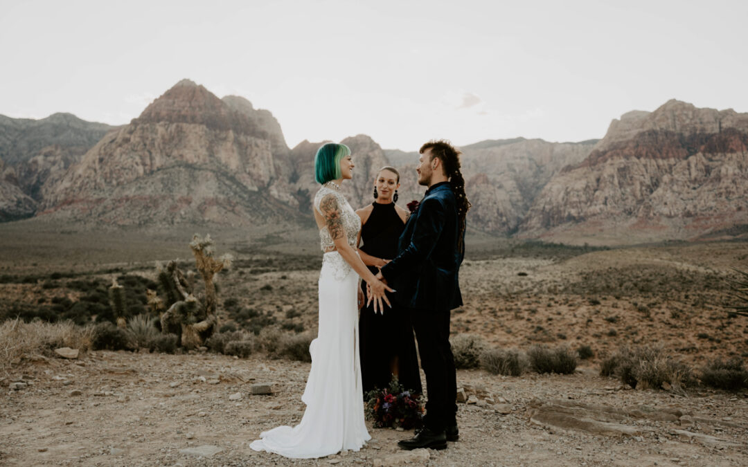 A bride with blue hair stands with her groom and a female officiant in a Vegas desert with mountains in the background.