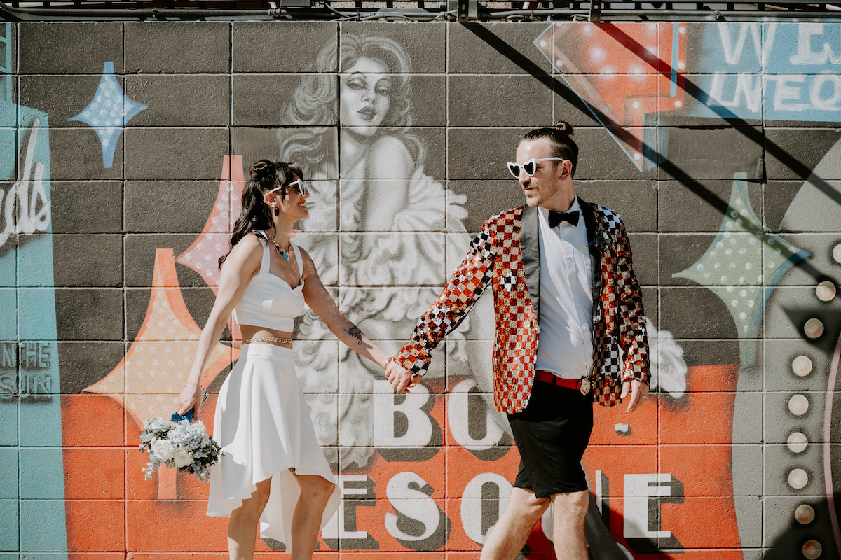 behind the scenes wedding content creation bride and groom in rock-n-roll attire holding hands in front of a mural